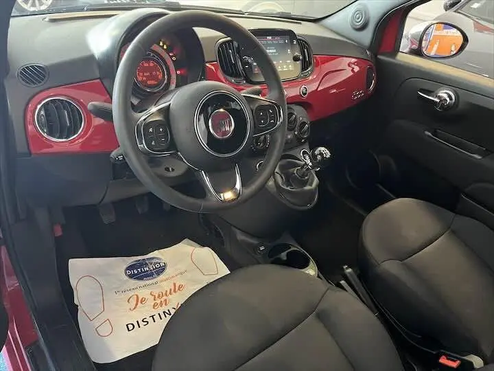 Vue intérieure côté conducteur de la Fiat 500 rouge 2023, tableau de bord brillant et volant multifonction avec boîte manuelle.