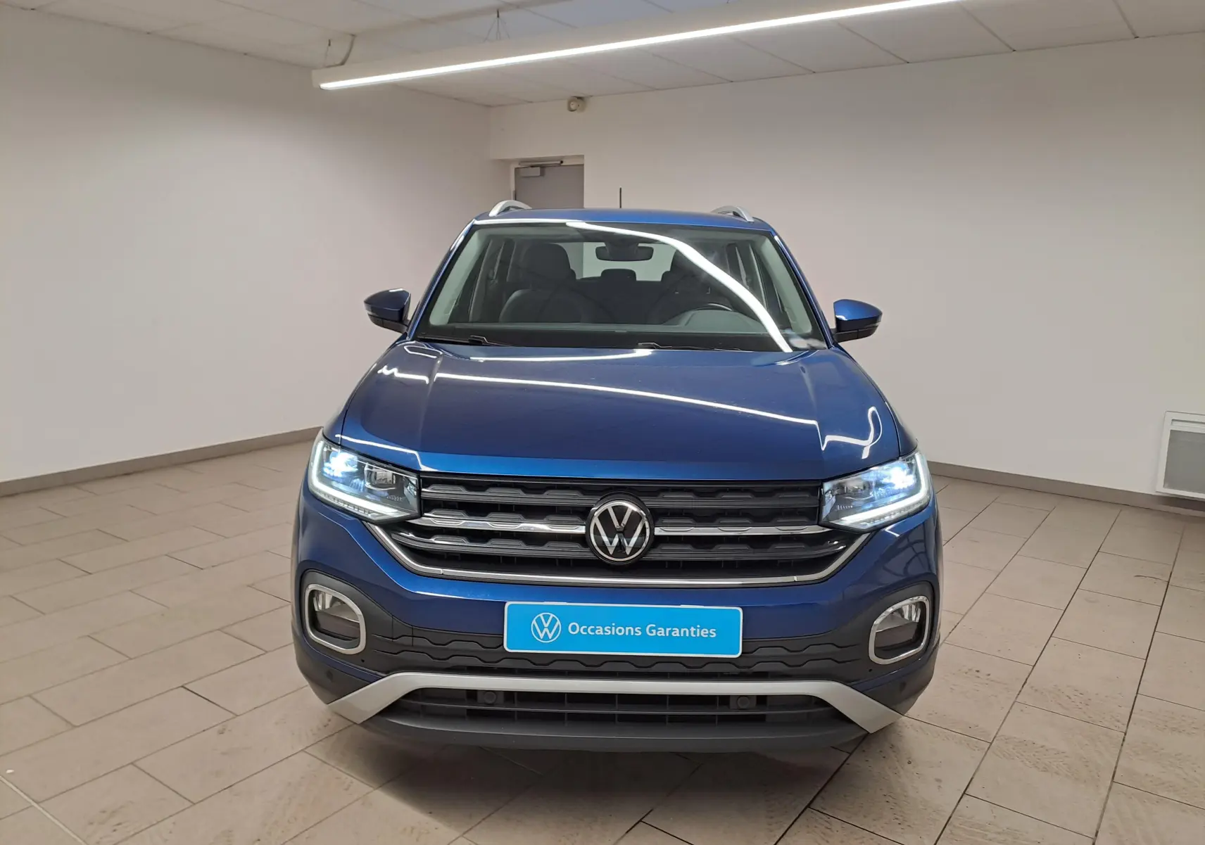Vue frontale d'un Volkswagen T-Cross 1.0 TSI 110 DSG7 Style bleu récif avec phares allumés en intérieur.