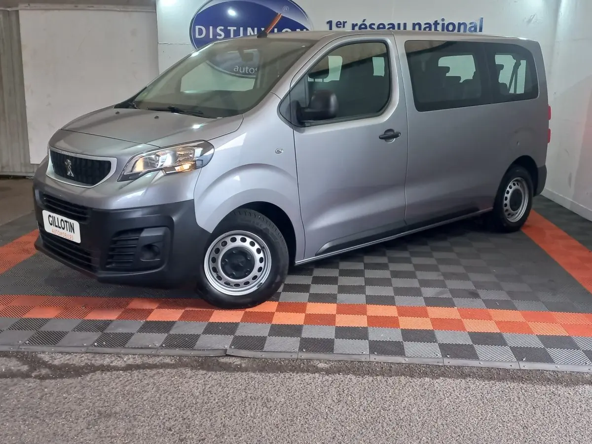Vue 3/4 avant droit d’un Peugeot Traveller gris clair avec jantes acier et calandre noire, stationné en intérieur.