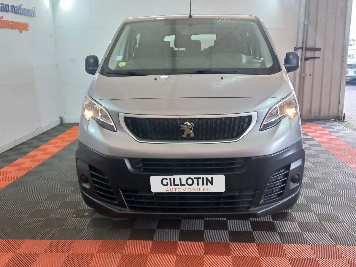 Vue frontale d’un Peugeot Traveller gris clair 2021 avec phares allumés et calandre noire, en intérieur showroom.