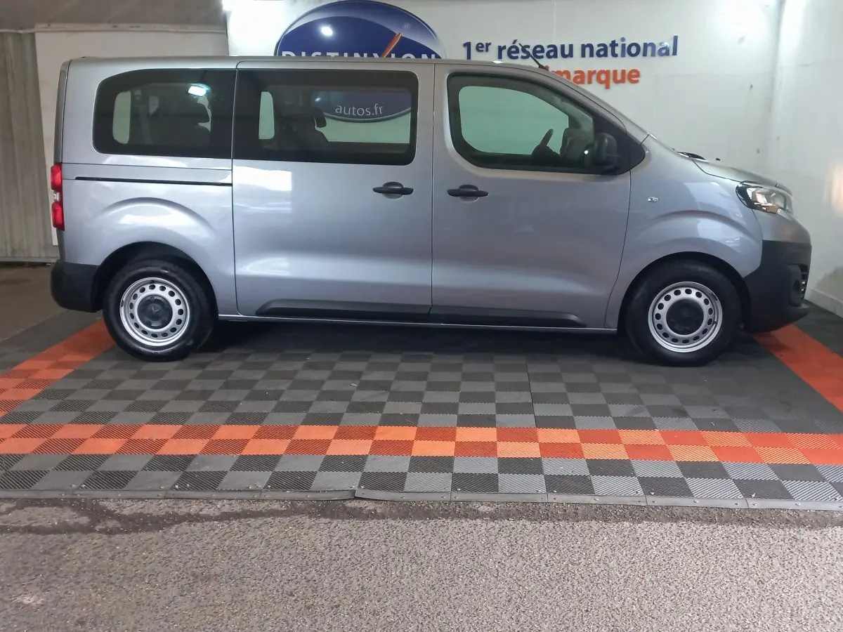 Vue de profil côté droit d’un Peugeot Traveller gris clair 2021 avec vitres arrière teintées et jantes acier simples.