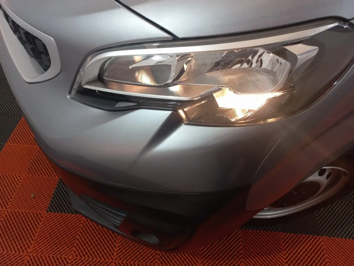 Gros plan sur le phare avant droit du Peugeot Traveller gris clair, avec partie de la calandre et roue visible.