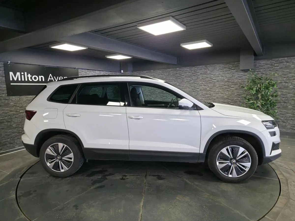 Vue de profil côté gauche d'un Skoda Karoq blanc 2023 avec jantes alliage et barres de toit noires en intérieur showroom.