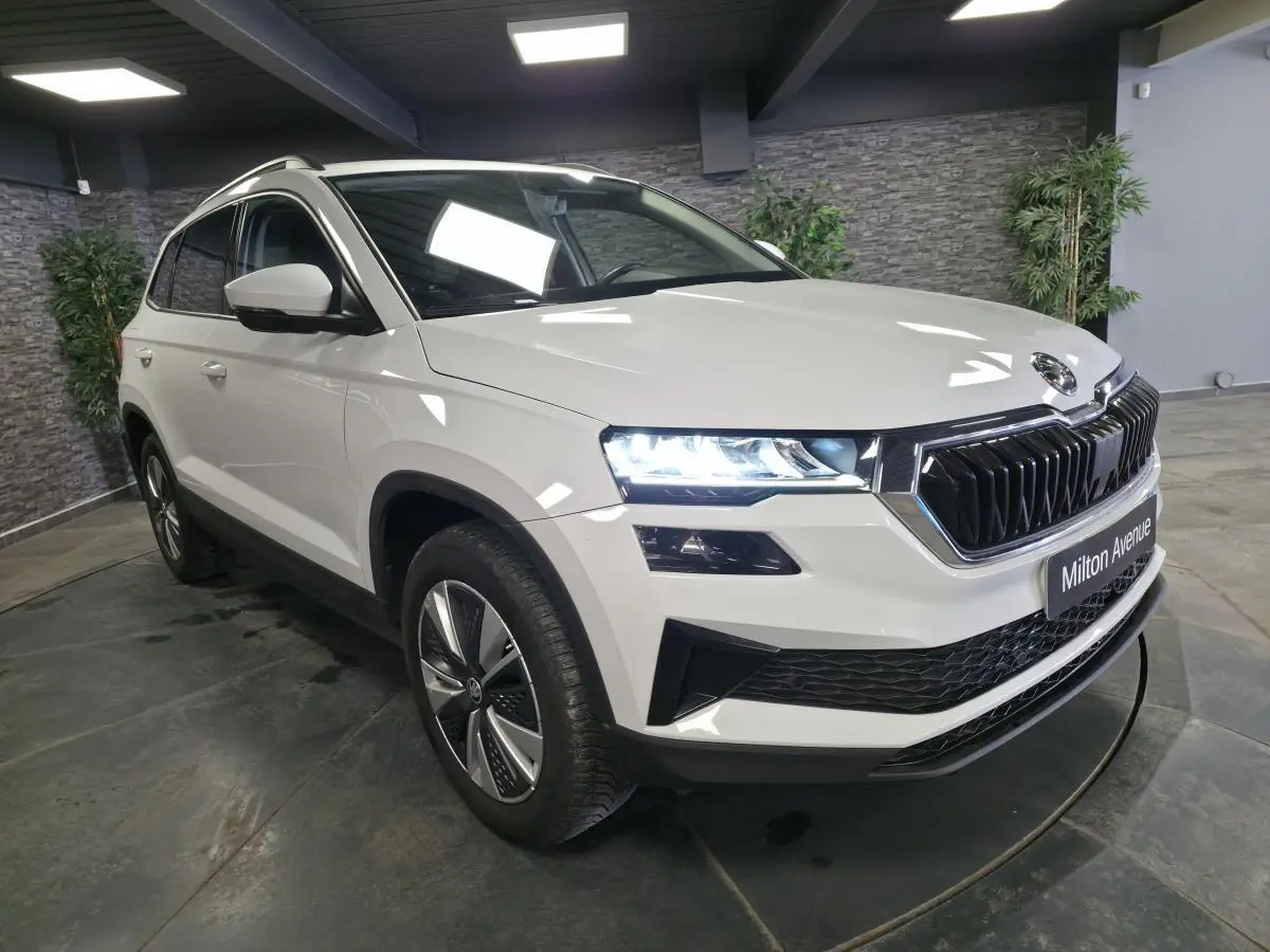 Vue 3/4 avant droit d'un Skoda Karoq blanc avec calandre noire et phares allumés en intérieur showroom.