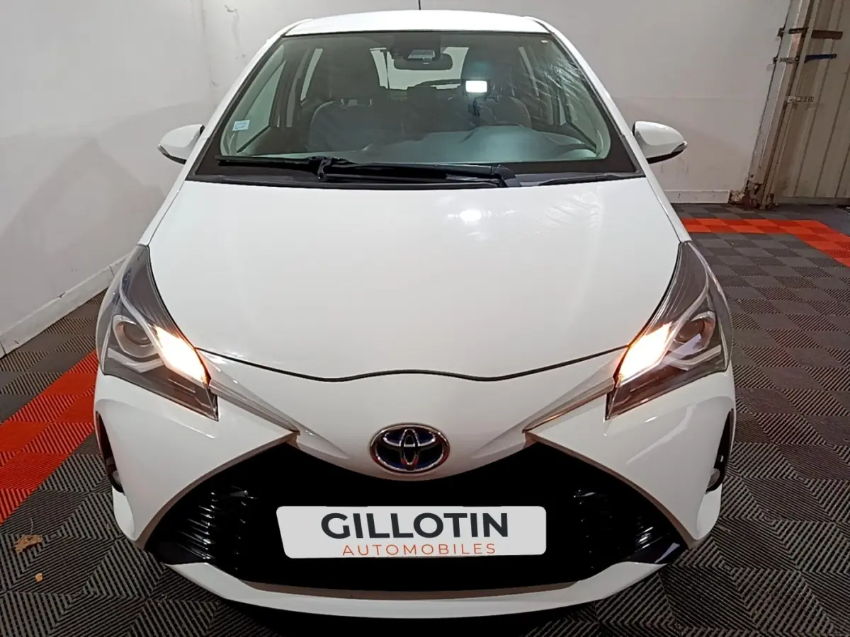 Vue avant d'une Toyota Yaris blanche 2019 avec phares allumés dans un garage au sol carrelé noir et rouge.