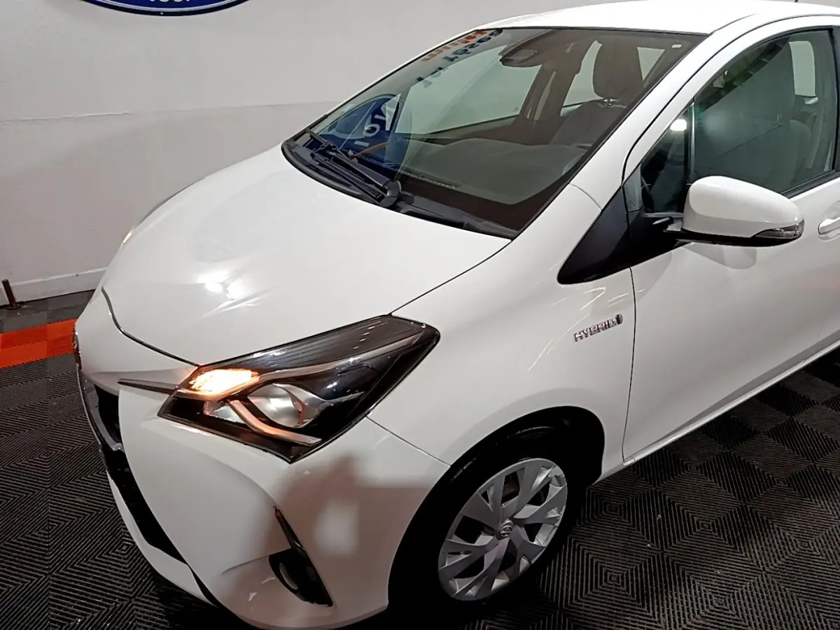 Toyota Yaris hybride blanche vue en 3/4 avant droit avec phares allumés et badge Hybrid sur l'aile avant.