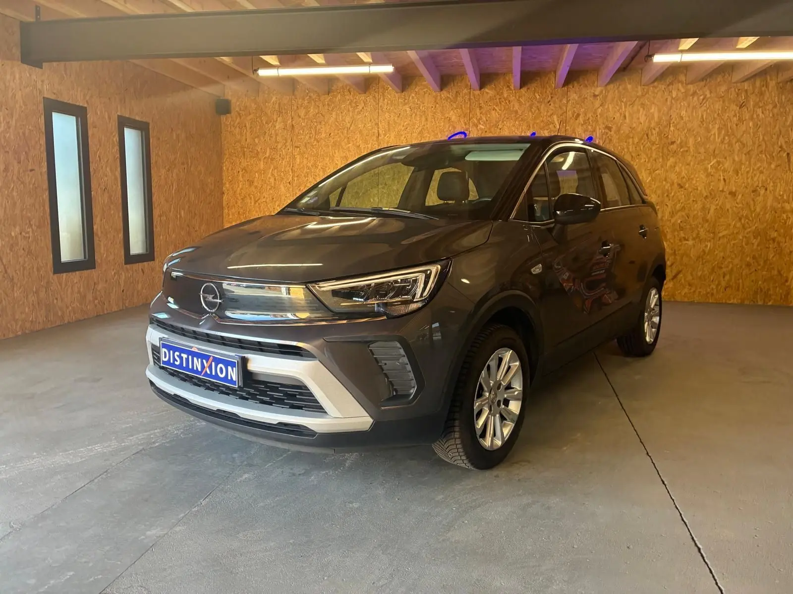 Opel Crossland gris en 3/4 avant droit, avec calandre chromée et jantes alliage dans un garage en bois.