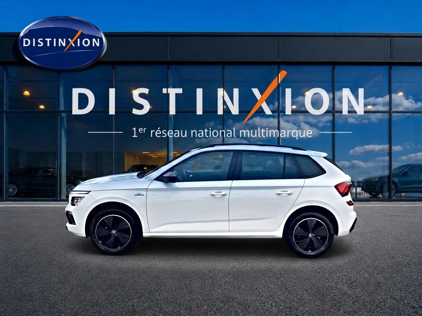 Profil côté gauche d'un Skoda Kamiq blanc nacré Monte Carlo avec jantes noires devant une vitrine Distinxion.