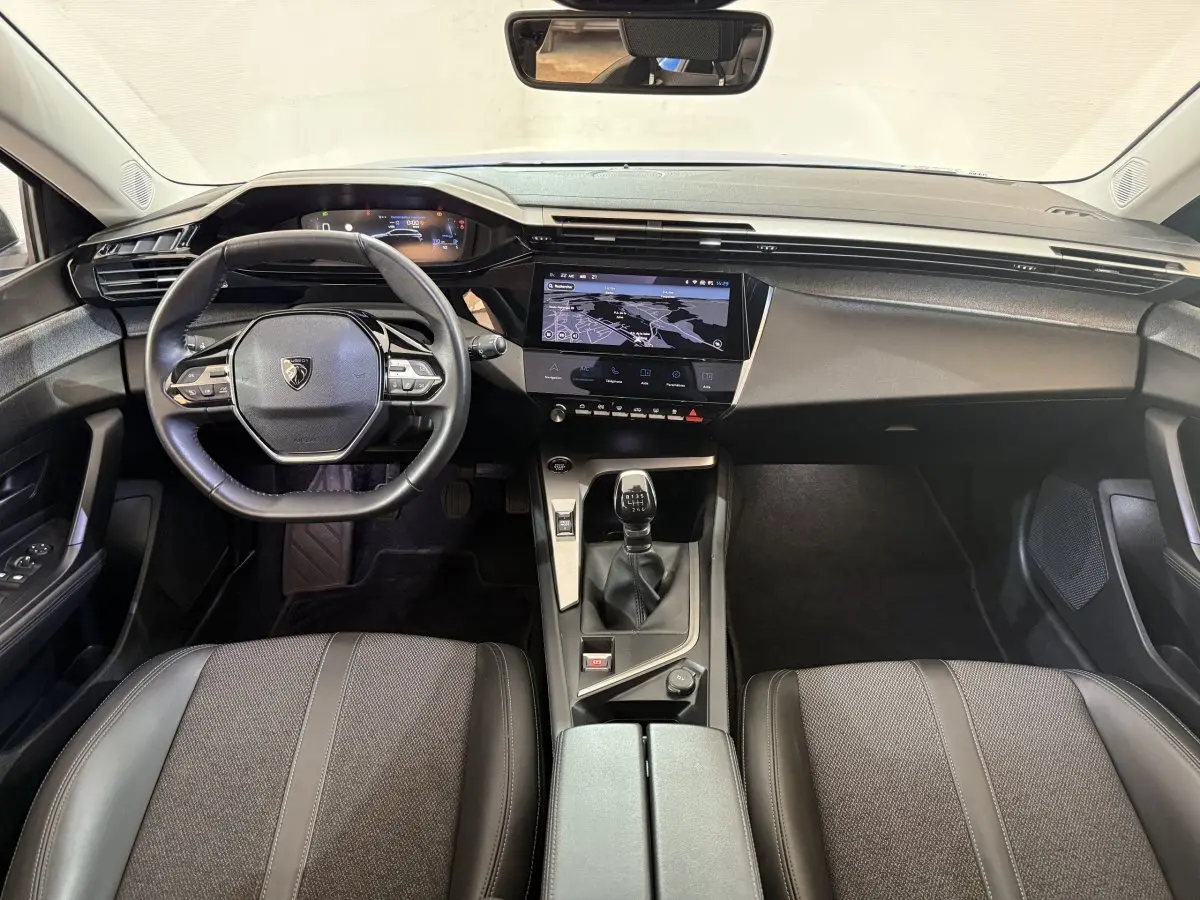 Vue intérieure avant de la Peugeot 308 2023, tableau de bord noir avec écran tactile central et volant cuir multifonctions.