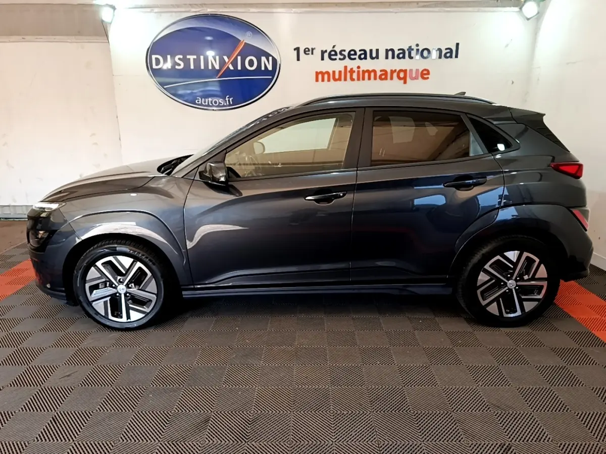 Vue de profil côté gauche d'un Hyundai Kona électrique gris foncé avec jantes distinctives en intérieur showroom.