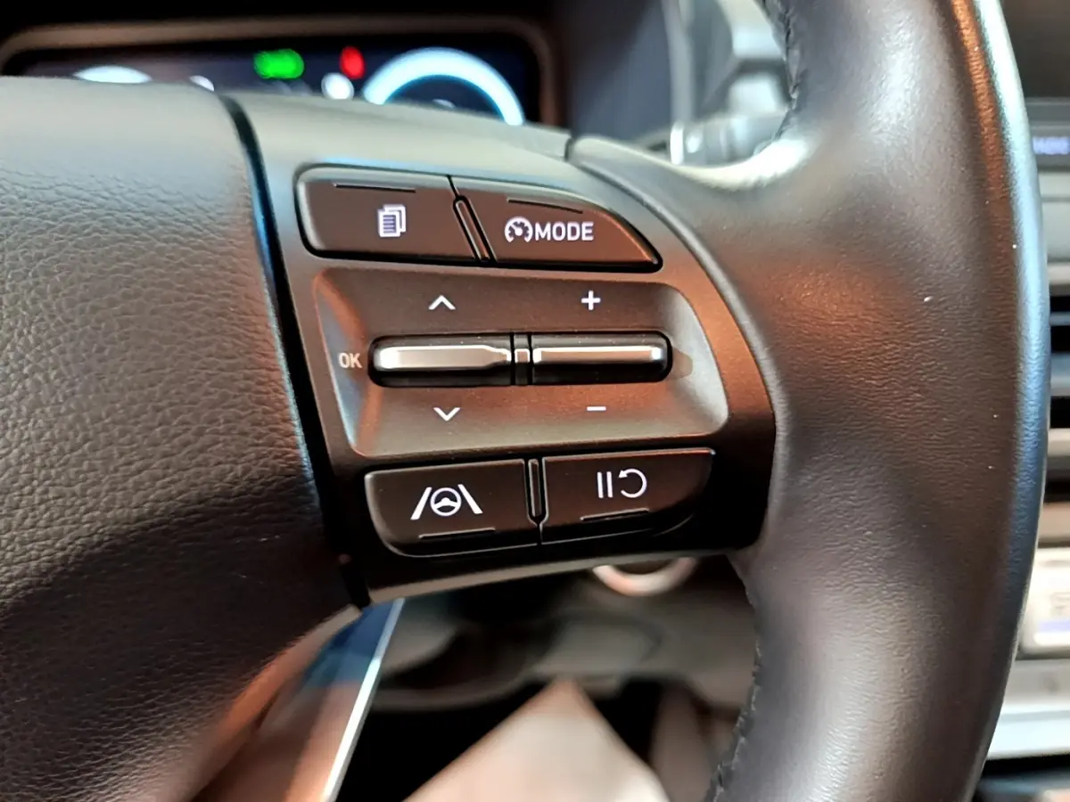 Gros plan sur les commandes du volant cuir noir du Hyundai Kona électrique, avec boutons multifonctions et régulateur de vitesse.
