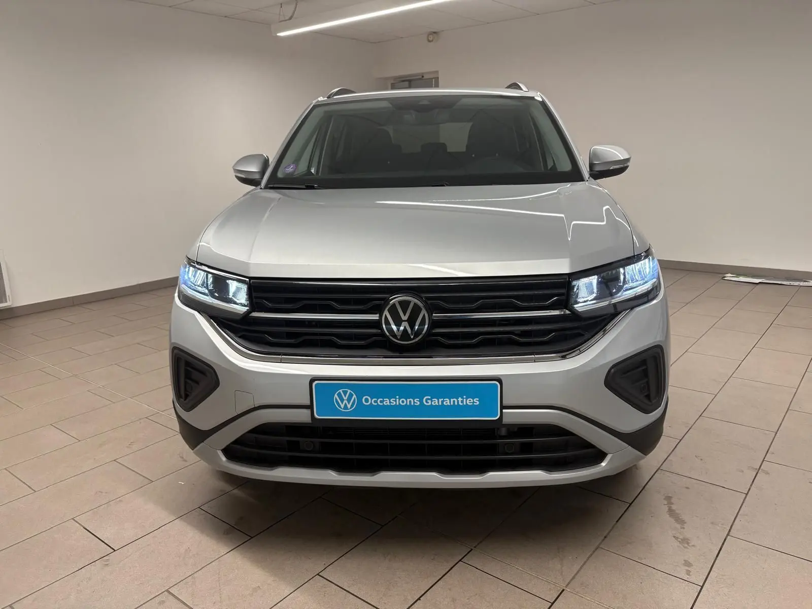 Vue frontale d'un Volkswagen T-Cross 2024 en reflet d'argent, avec phares LED allumés et plaque "Occasions Garantis".