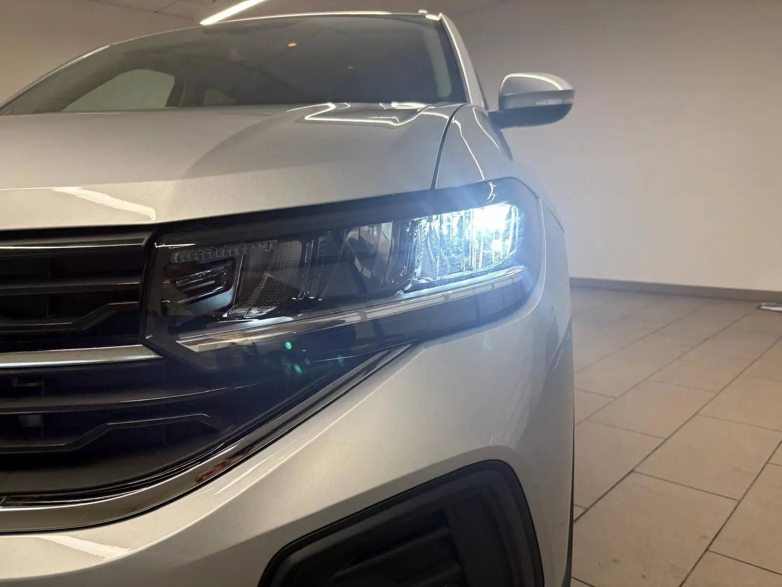 Gros plan sur le phare avant droit allumé d'un Volkswagen T-Cross 2024 couleur reflet d'argent, intérieur showroom.