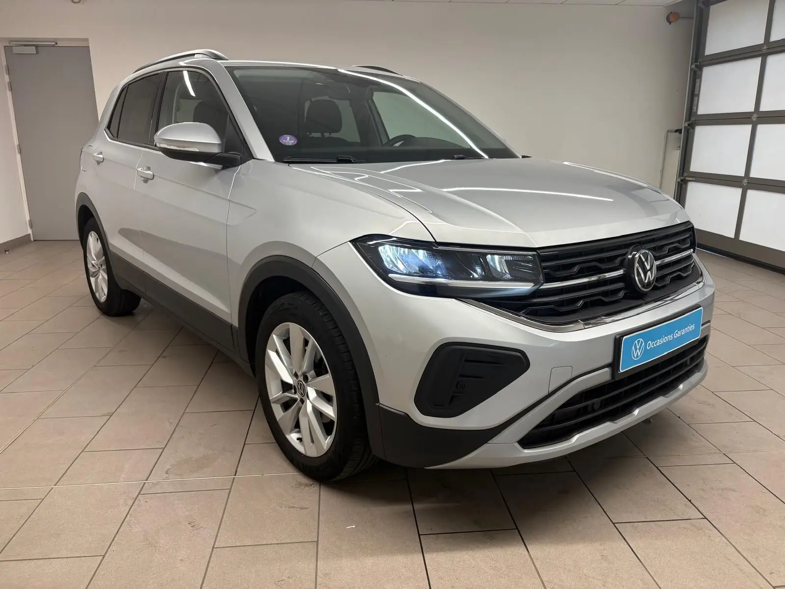 Volkswagen T-Cross 2024 argent reflet, vue 3/4 avant droit en intérieur avec phares LED allumés.