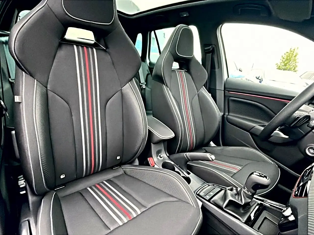 Vue intérieure avant du Skoda Kamiq 2026 avec sièges sport noirs à bandes rouges et grises, volant multifonction et levier de vitesse.
