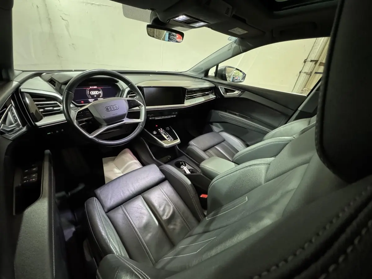 Intérieur noir de l'Audi Q4 E-TRON 50 quattro 2022, vue depuis l'arrière côté conducteur sur le tableau de bord et volant cuir multifonctions.