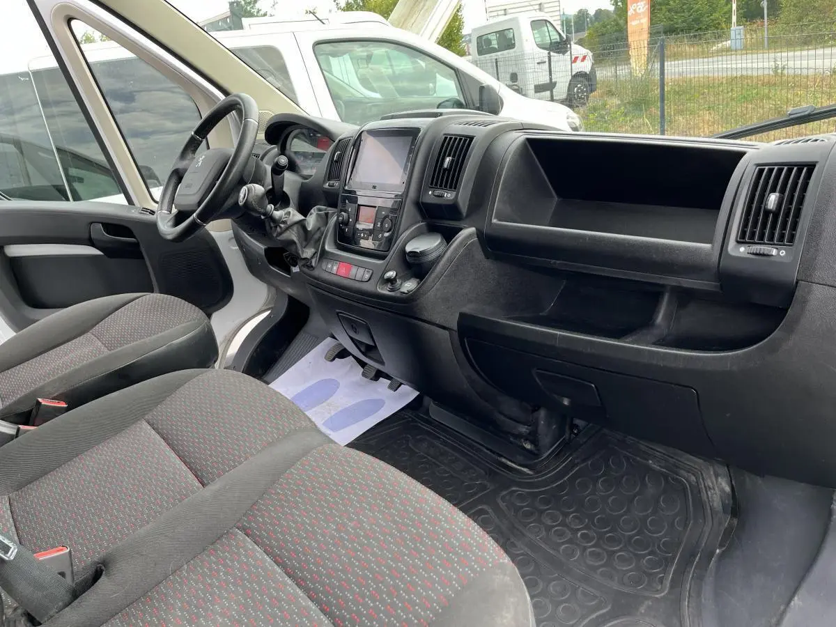Vue intérieure côté conducteur du Peugeot Boxer Fourgon 2023, montrant tableau de bord noir et sièges tissu gris à motifs rouges.