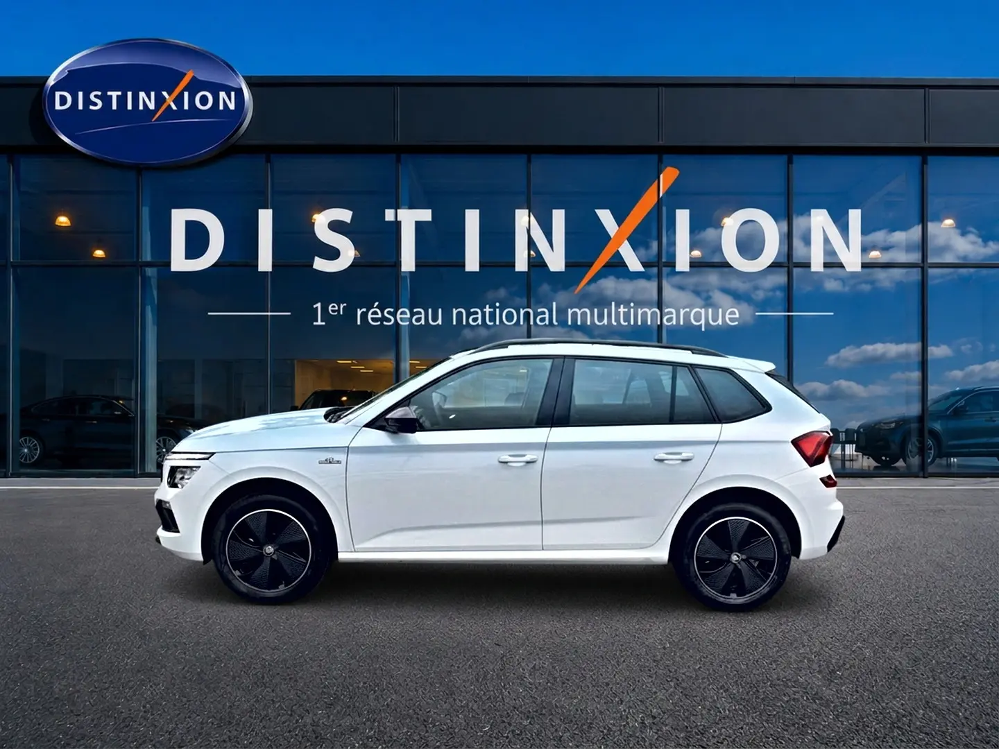 Vue latérale droite d'un Skoda Kamiq blanc nacré avec jantes noires devant un bâtiment vitré Distinxion.