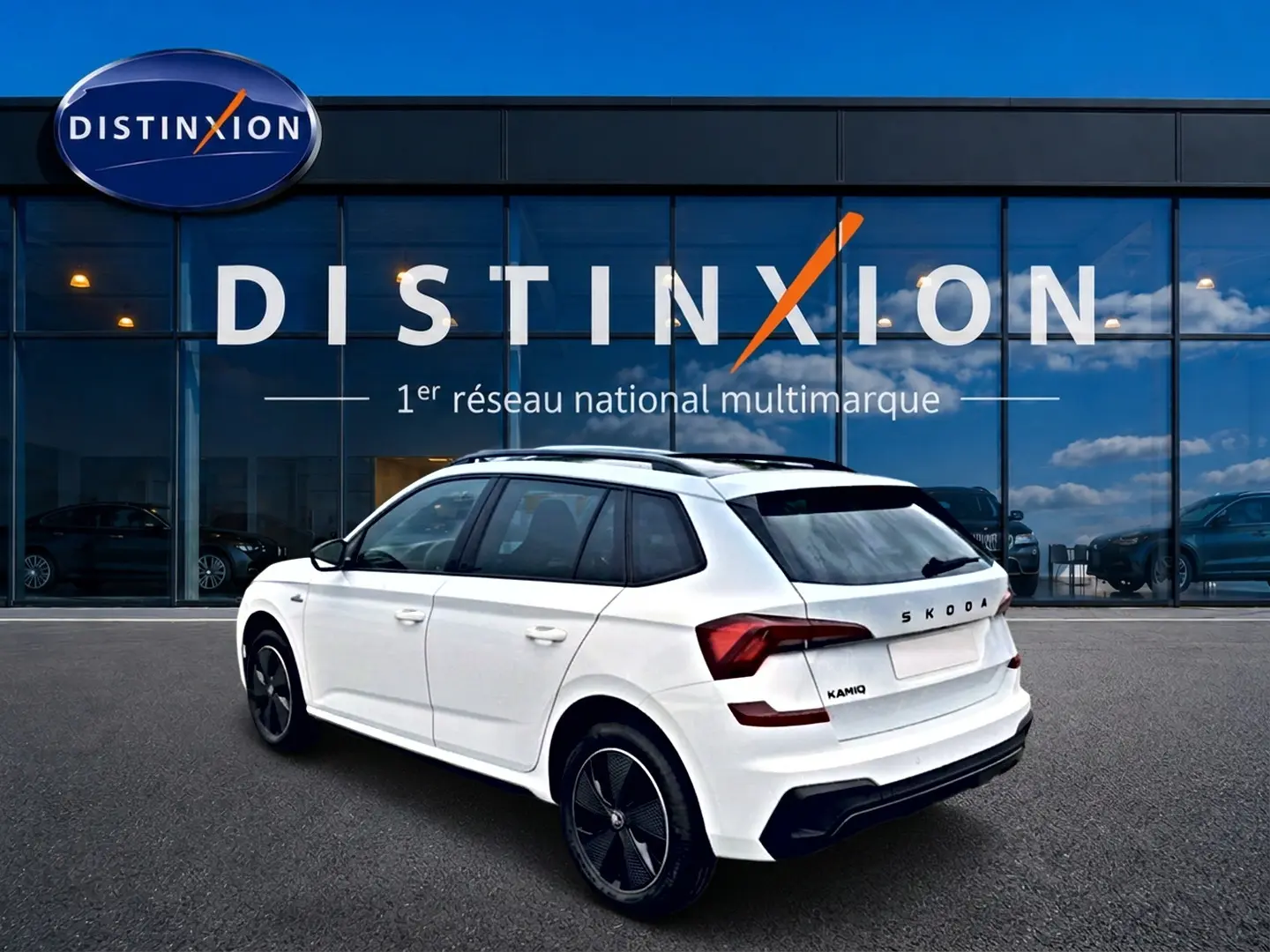 Vue 3/4 arrière droite d’un Skoda Kamiq blanc nacré avec jantes noires devant un bâtiment vitré Distinxion.
