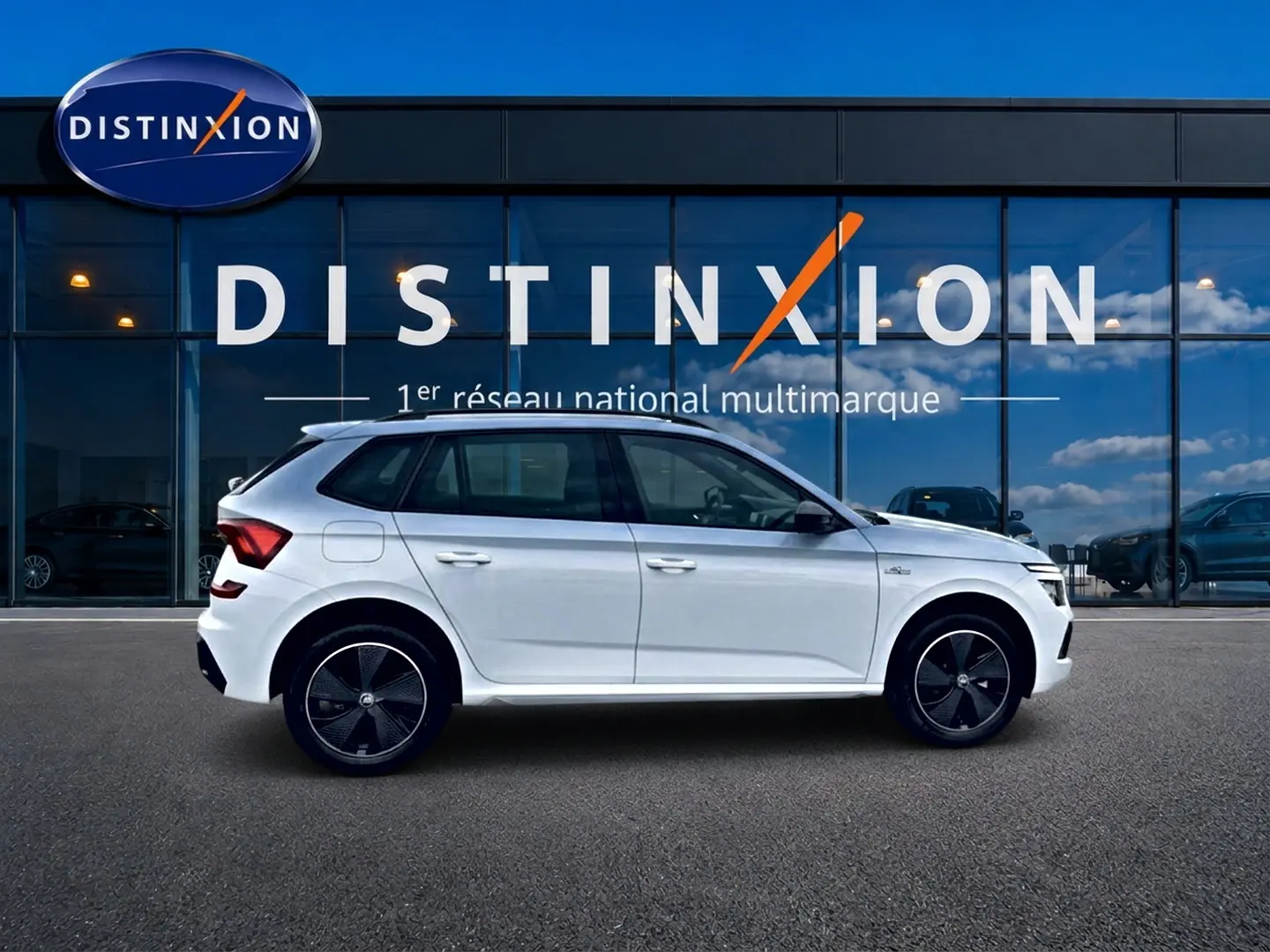 Vue de profil côté gauche d'un Skoda Kamiq blanc nacré avec jantes noires devant une vitrine Distinxion.