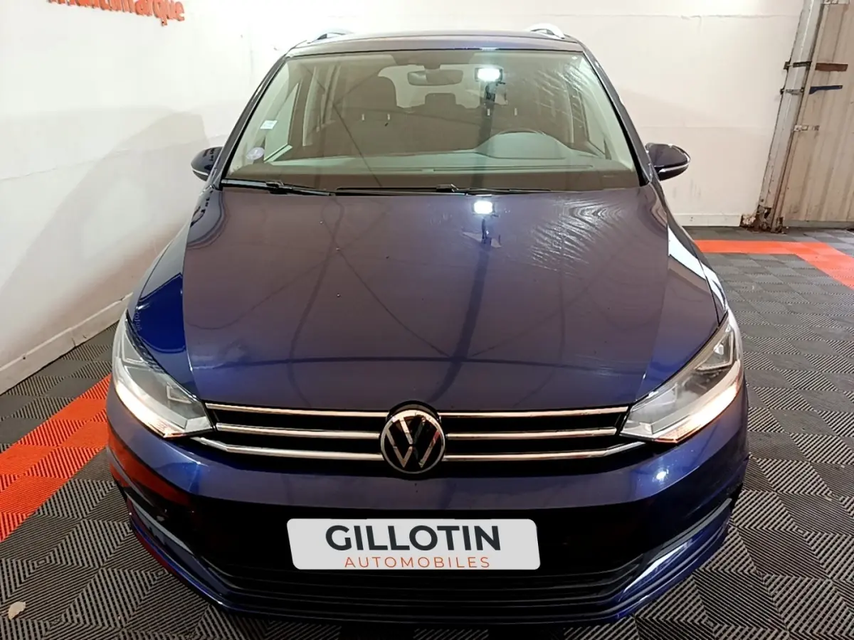 Vue avant d'un Volkswagen Touran bleu 2021 avec calandre chromée et phares allumés dans un garage.