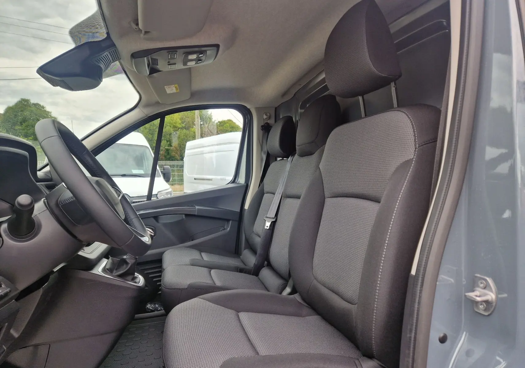 Vue intérieure côté conducteur du Renault Trafic Fourgon gris Highland, montrant les sièges avant et la banquette passager avec bureau mobile.