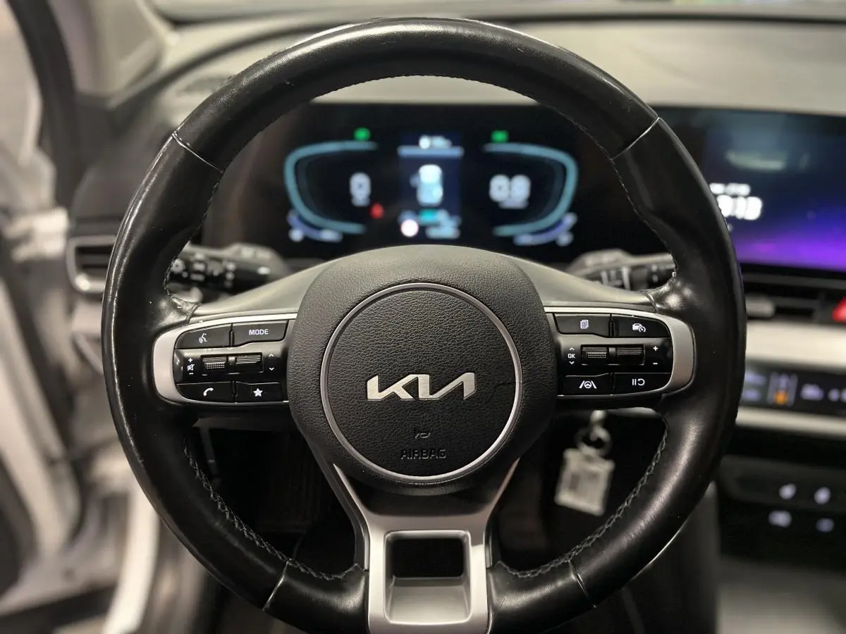 Gros plan sur le volant cuir noir du Kia Sportage 2022 avec commandes intégrées et tableau de bord digital flou en arrière-plan.