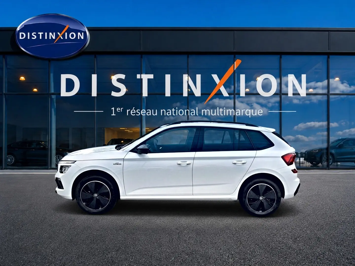 Vue de profil côté gauche d'un Skoda Kamiq blanc nacré avec jantes noires devant une vitrine Distinxion.