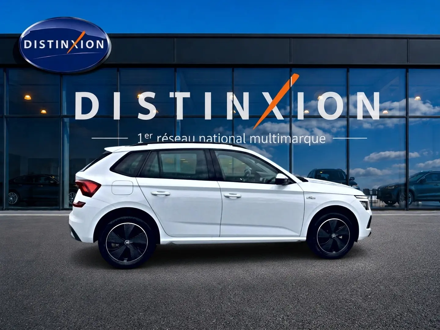 Vue latérale droite d'un Skoda Kamiq blanc nacré avec jantes noires devant une vitrine Distinxion.