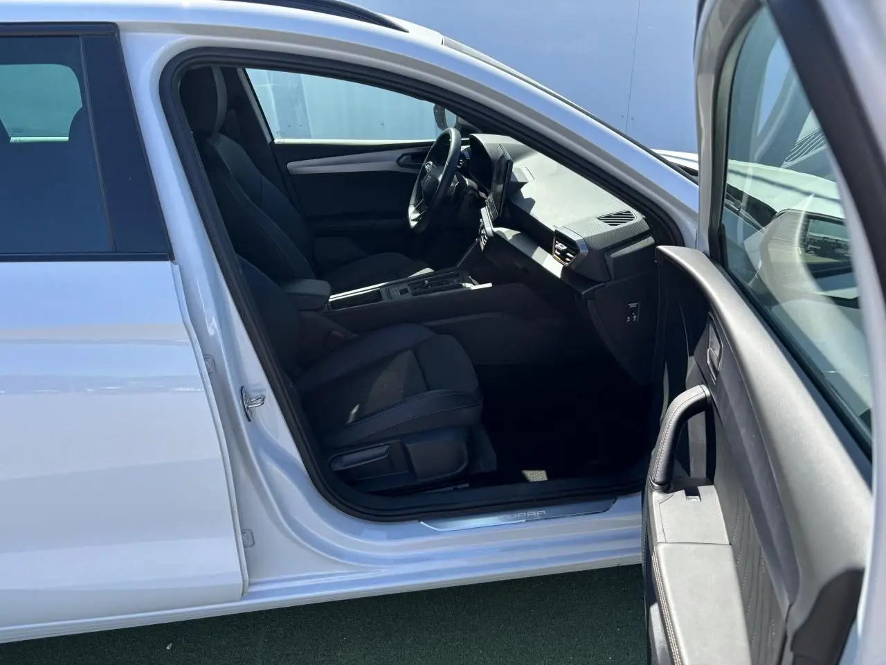 Vue intérieure côté conducteur d'une CUPRA Leon Sportstourer 2024 blanche Nevada, montrant sièges noirs et tableau de bord moderne.