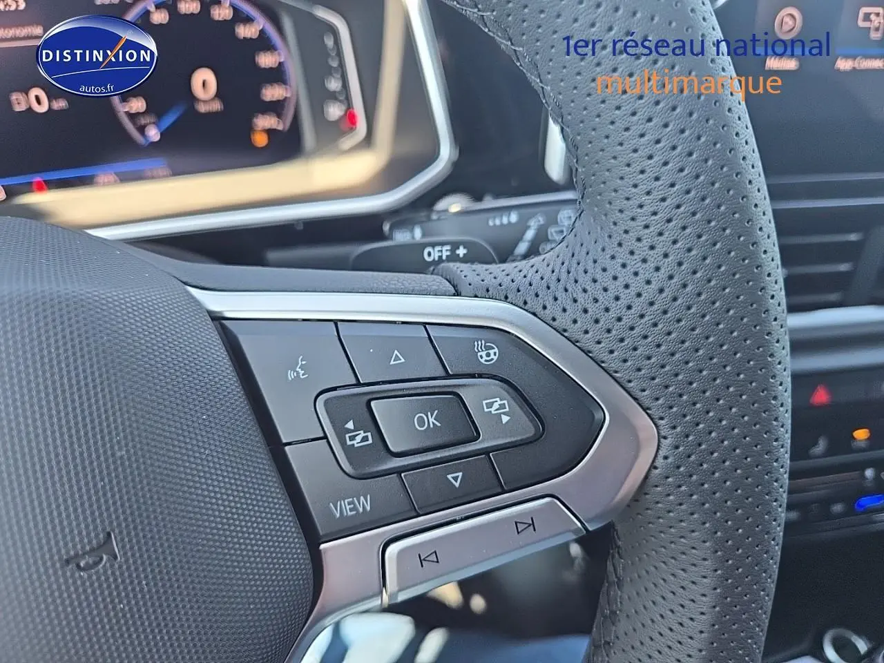 Gros plan sur le volant cuir perforé du Volkswagen T-Roc noir perle, avec commandes multifonctions et tableau de bord numérique flou.