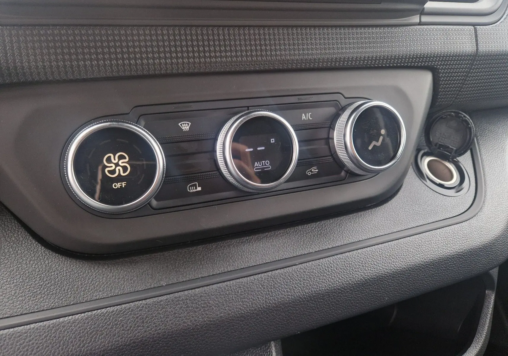 Gros plan sur les commandes de climatisation du Renault Trafic Fourgon gris urbain, avec boutons ronds et prise allume-cigare ouverte.