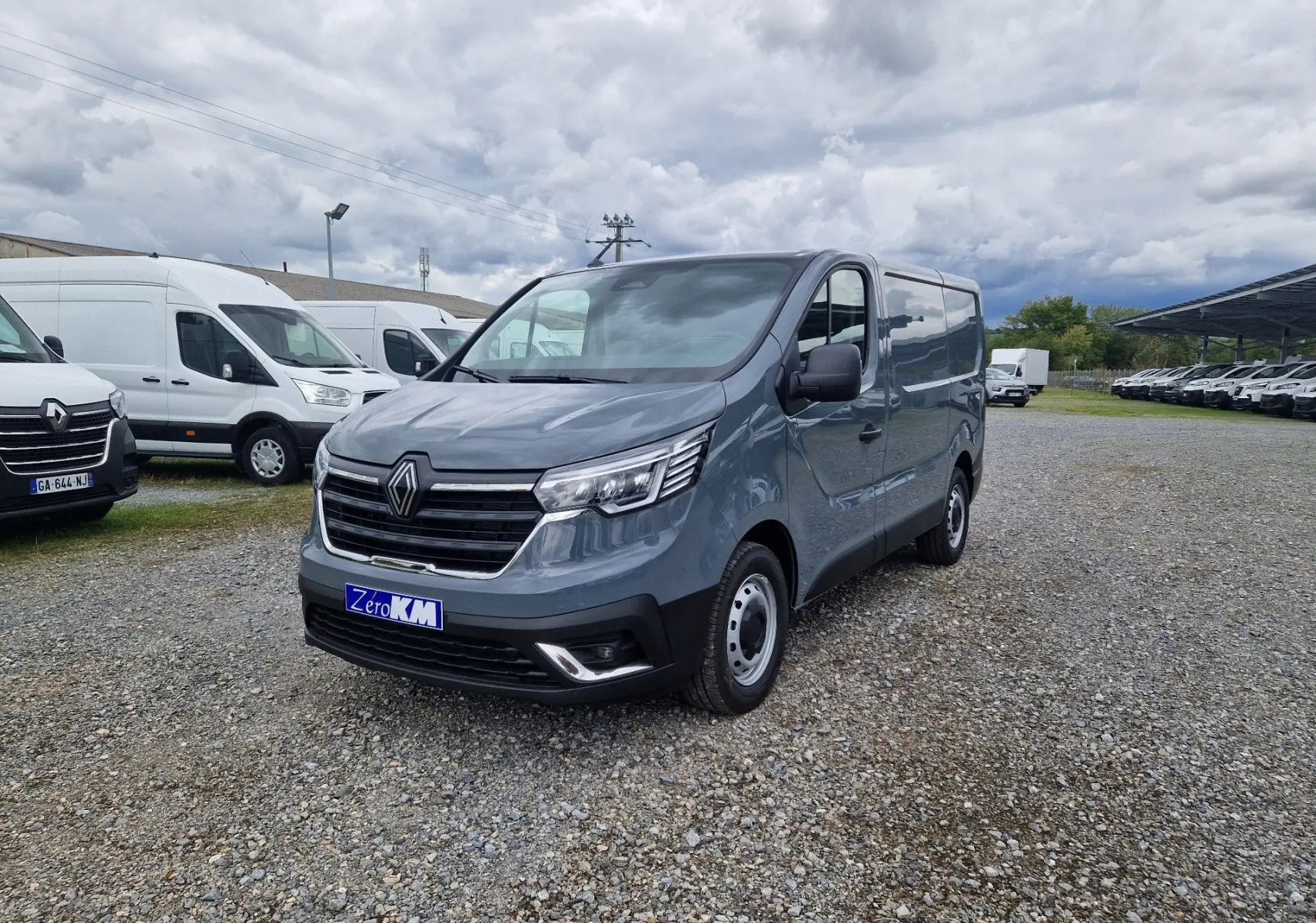 Renault Trafic Fourgon gris urbain vu en 3/4 avant droit sur un parking gravillonné, avec calandre noire et phares LED.