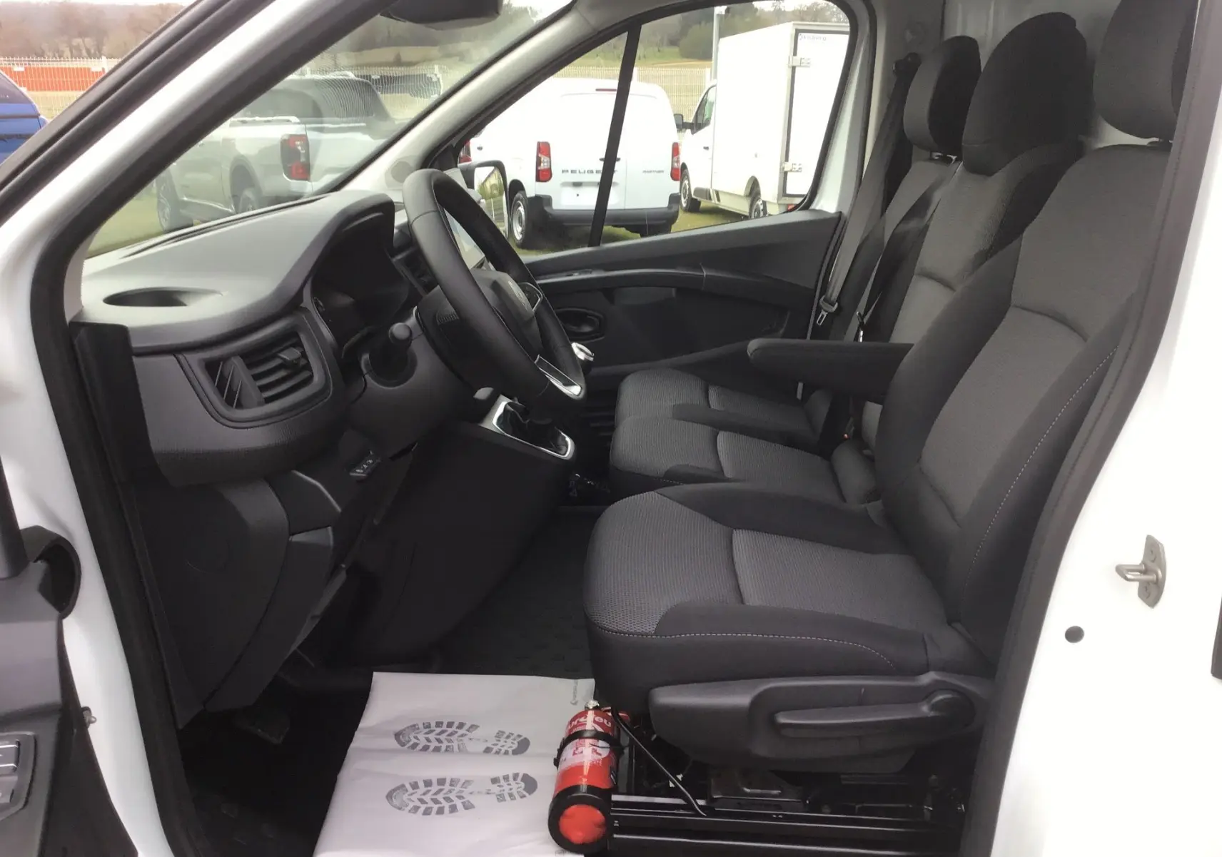 Vue intérieure côté conducteur du Renault Trafic blanc, montrant les sièges noirs et le tableau de bord avec volant et levier de vitesse.