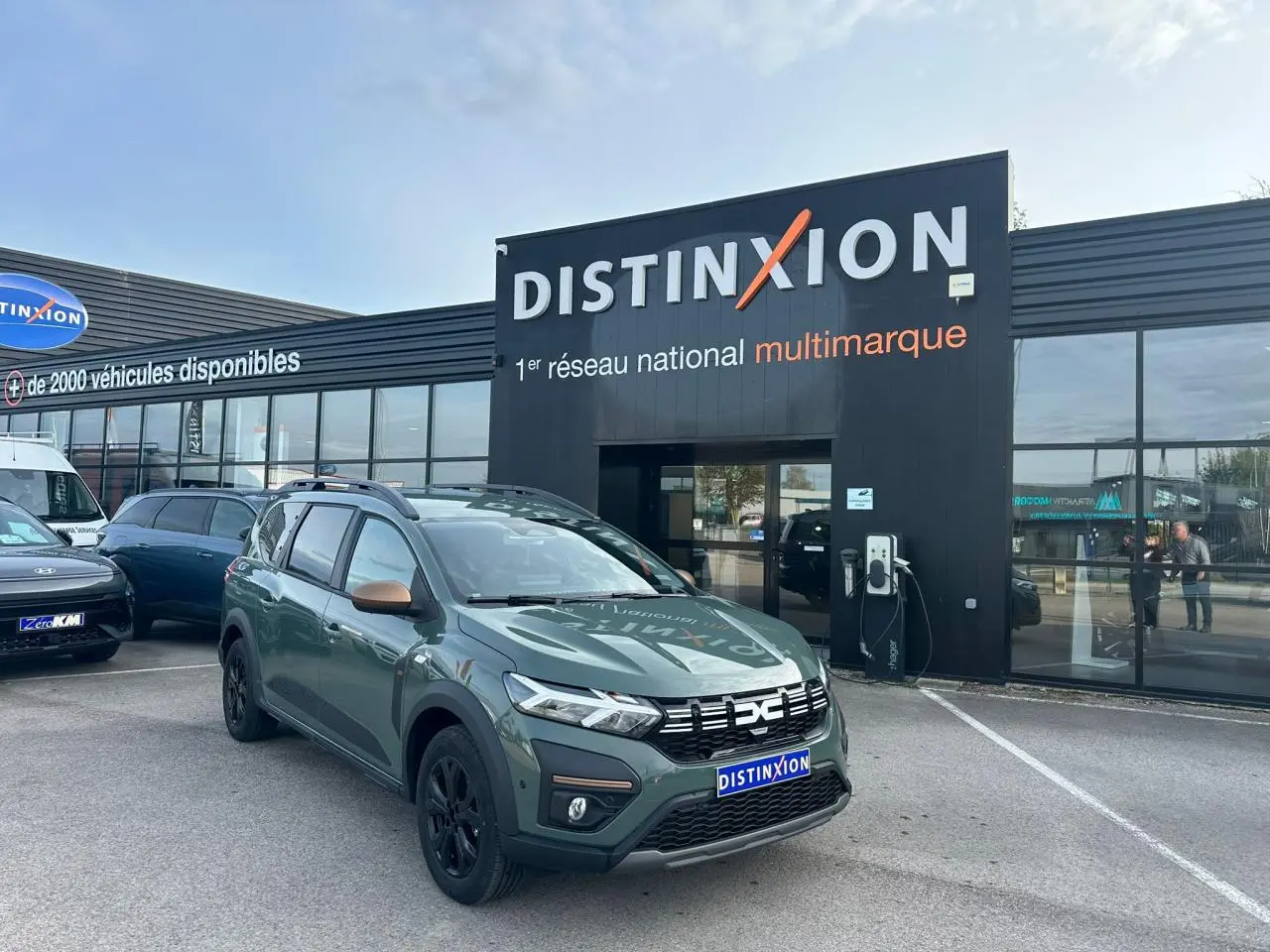 Vue 3/4 avant d'un Dacia Jogger kaki lichen métal avec calandre noire et jantes noires devant un concessionnaire Distinxion.