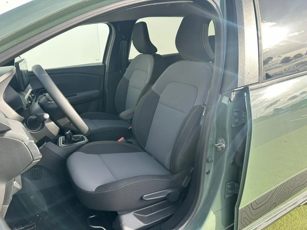Vue intérieure côté conducteur du Dacia Jogger kaki lichen, mettant en valeur les sièges avant gris et noirs avec surpiqûres contrastées.