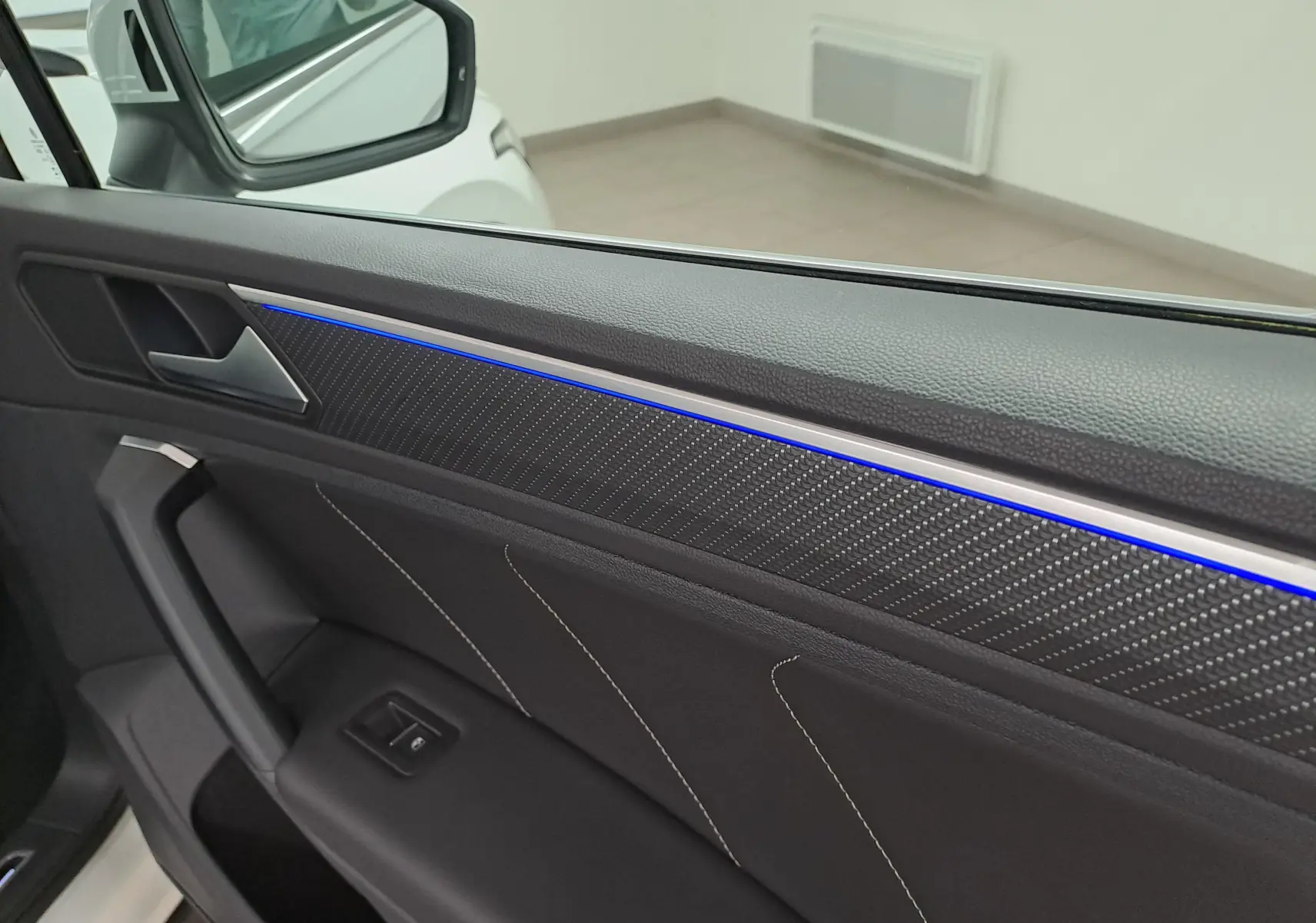 Vue intérieure de la porte avant gauche du Volkswagen Tiguan blanc, avec éclairage d'ambiance bleu et finition carbone.