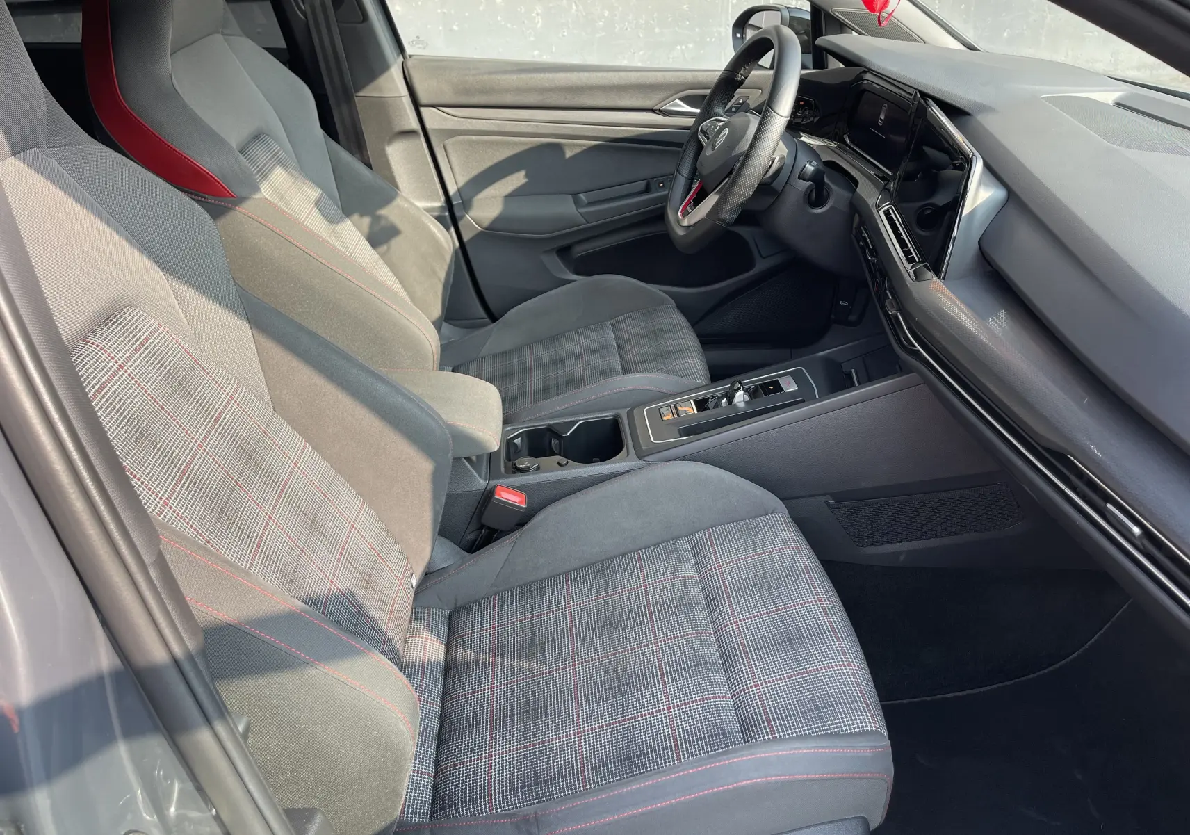 Intérieur côté conducteur de la Volkswagen Golf VIII GTI 2021, sièges tissu à motif écossais avec surpiqûres rouges et tableau de bord moderne.