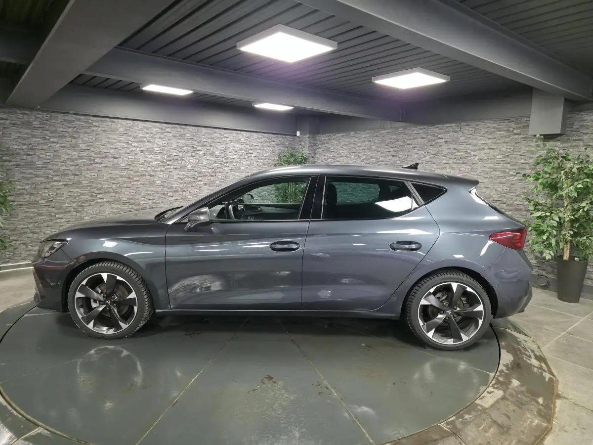 Vue de profil côté gauche d'une CUPRA Leon gris magnétique avec jantes alliage 18'' dans un showroom intérieur.
