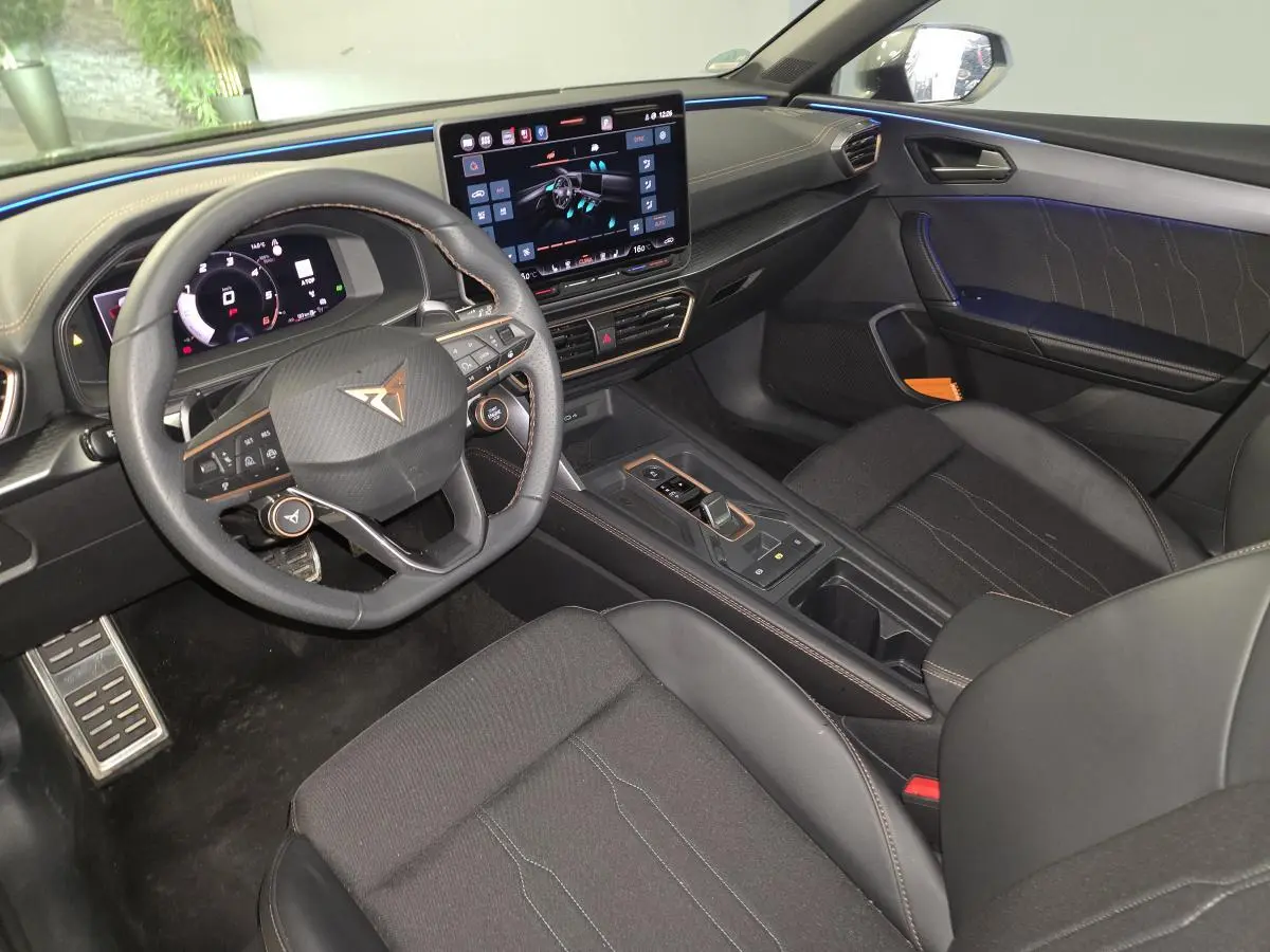 Intérieur avant de la CUPRA Leon 2025 gris, vue côté conducteur avec volant cuir, écran tactile et éclairage d’ambiance bleu.