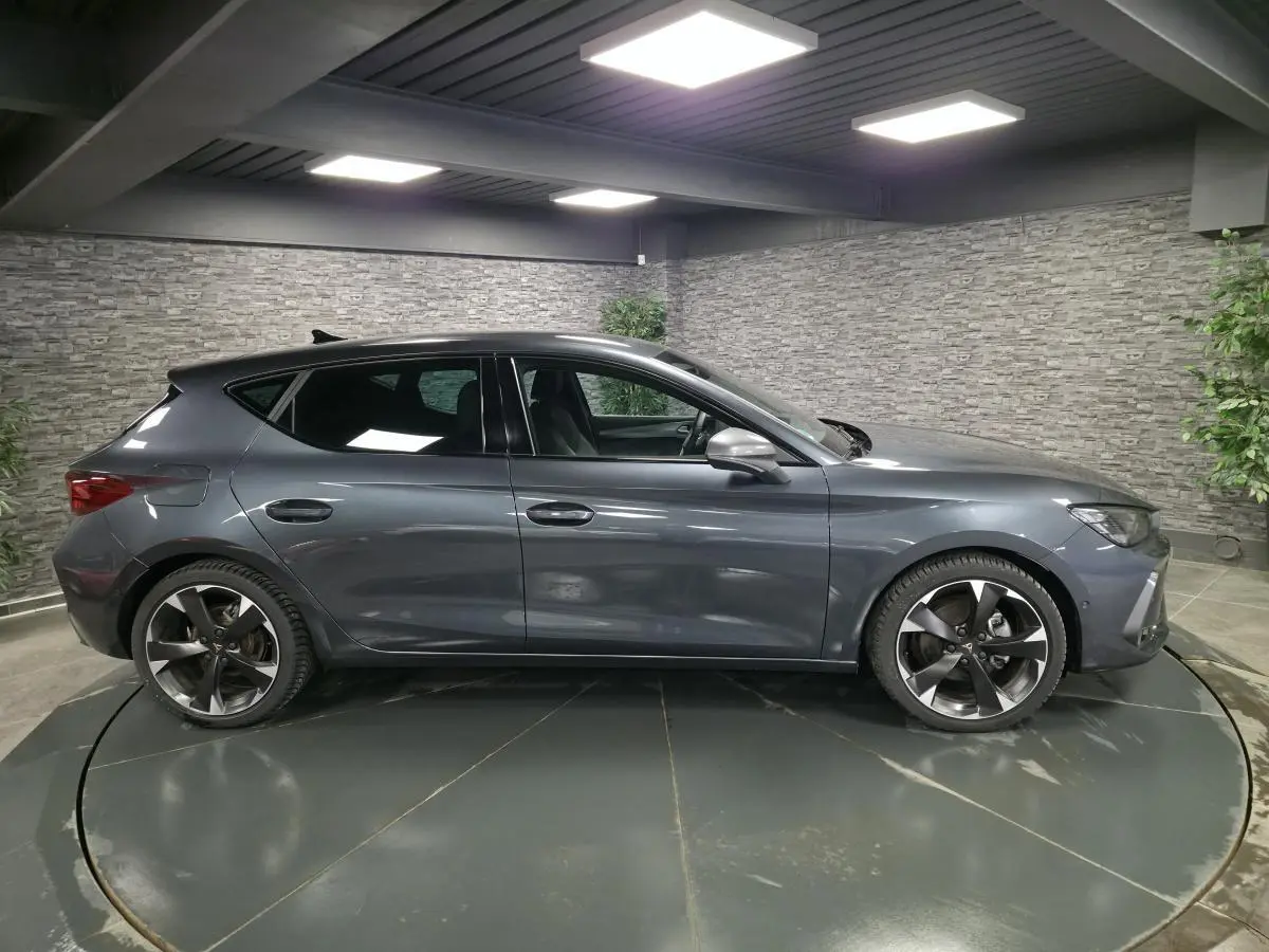 Vue de profil côté gauche d'une CUPRA Leon gris magnétique avec jantes alliage 18 pouces dans un showroom intérieur.