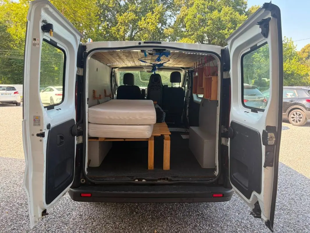 Vue arrière du Renault Trafic Fourgon blanc 2018, portes ouvertes montrant l’aménagement intérieur avec lit et rangements en bois.