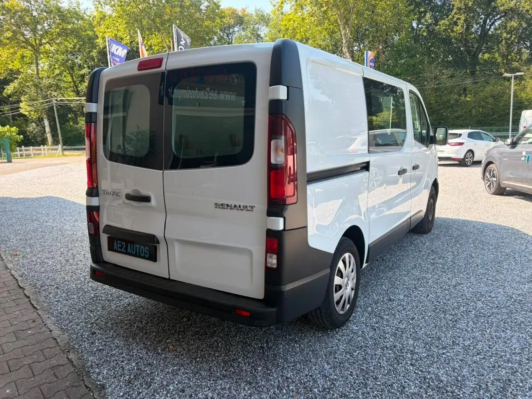 Vue 3/4 arrière droite d’un Renault Trafic Fourgon blanc de 2018 avec vitres arrière teintées et logo visible.