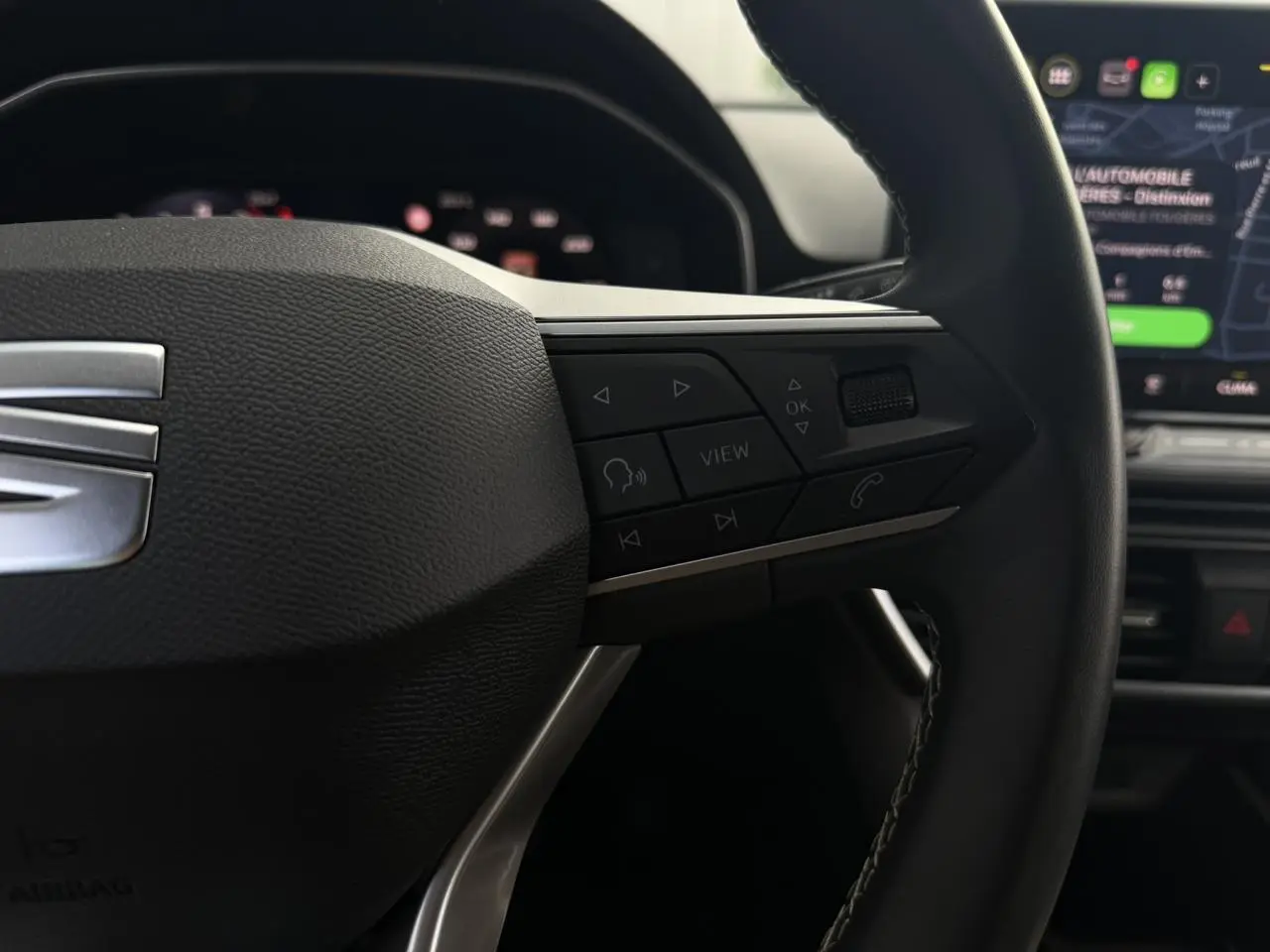 Gros plan sur le volant cuir multifonction noir du SEAT Leon ST 2025 avec commandes et écran tactile en arrière-plan flou.