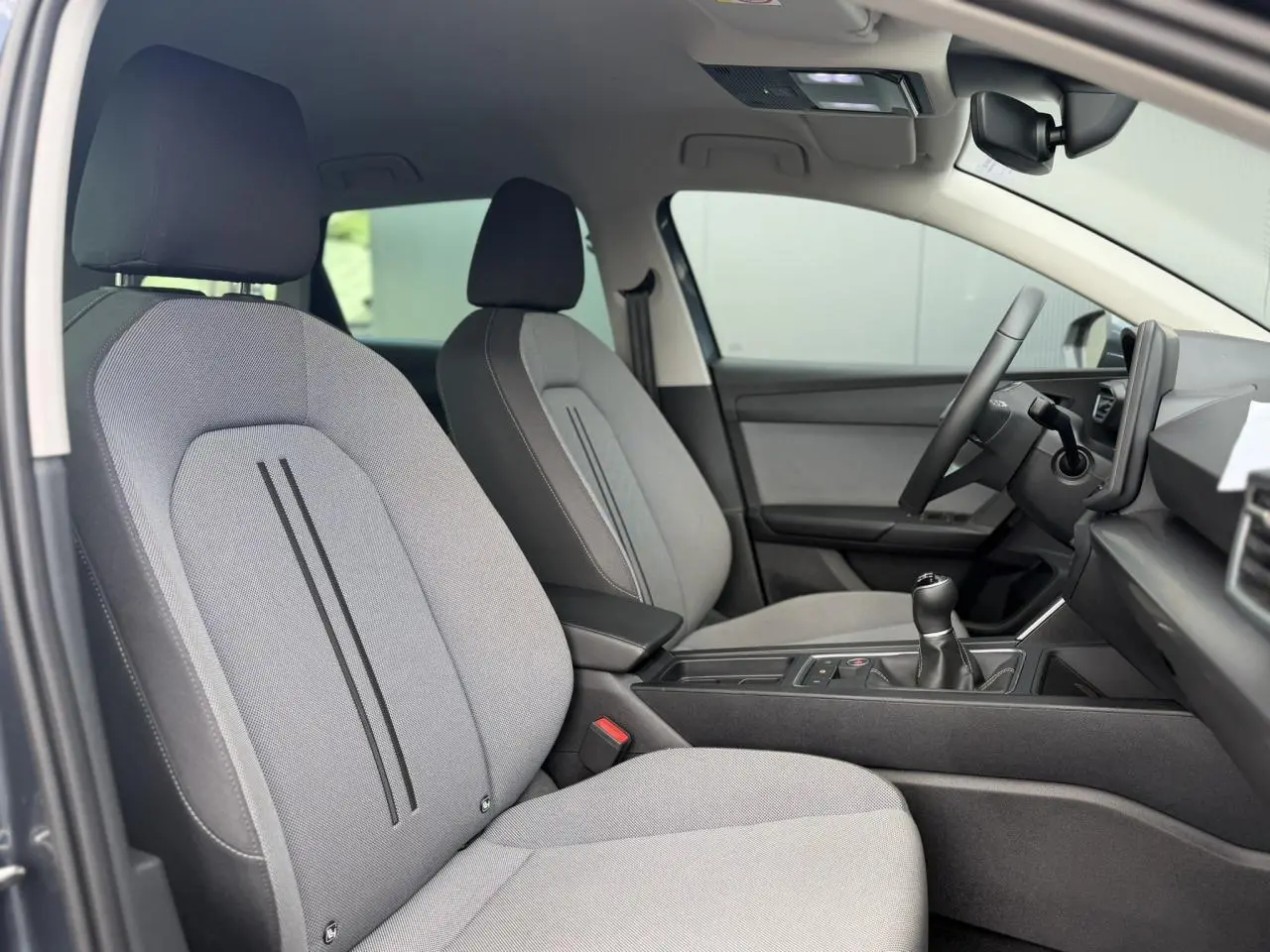 Vue intérieure côté conducteur de la SEAT LEON ST 2025, sièges textile gris clair avec console centrale et volant multifonction.