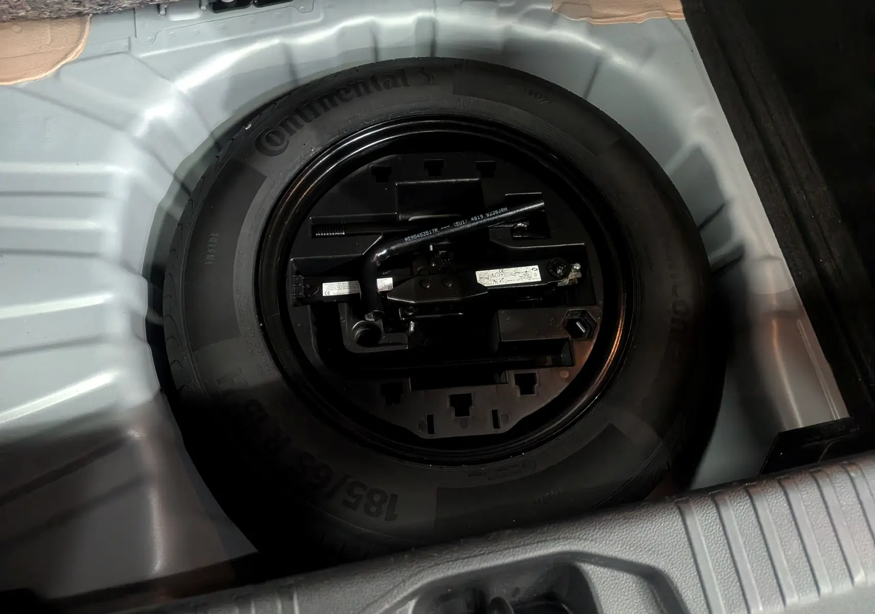 Vue plongeante sur la roue de secours noire et ses outils dans le coffre gris platine d'une Renault Clio V TCe 90 Auto-école.