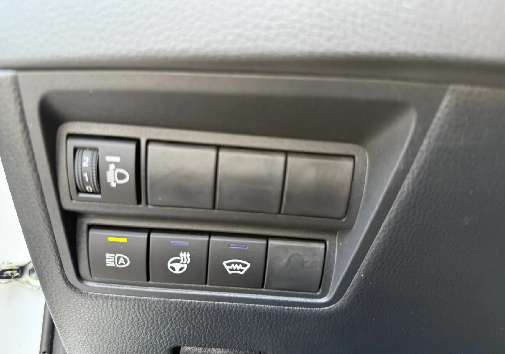 Gros plan sur les boutons de commande intérieurs noirs du Toyota Yaris Cross blanc, avec réglage des phares et chauffage du volant.