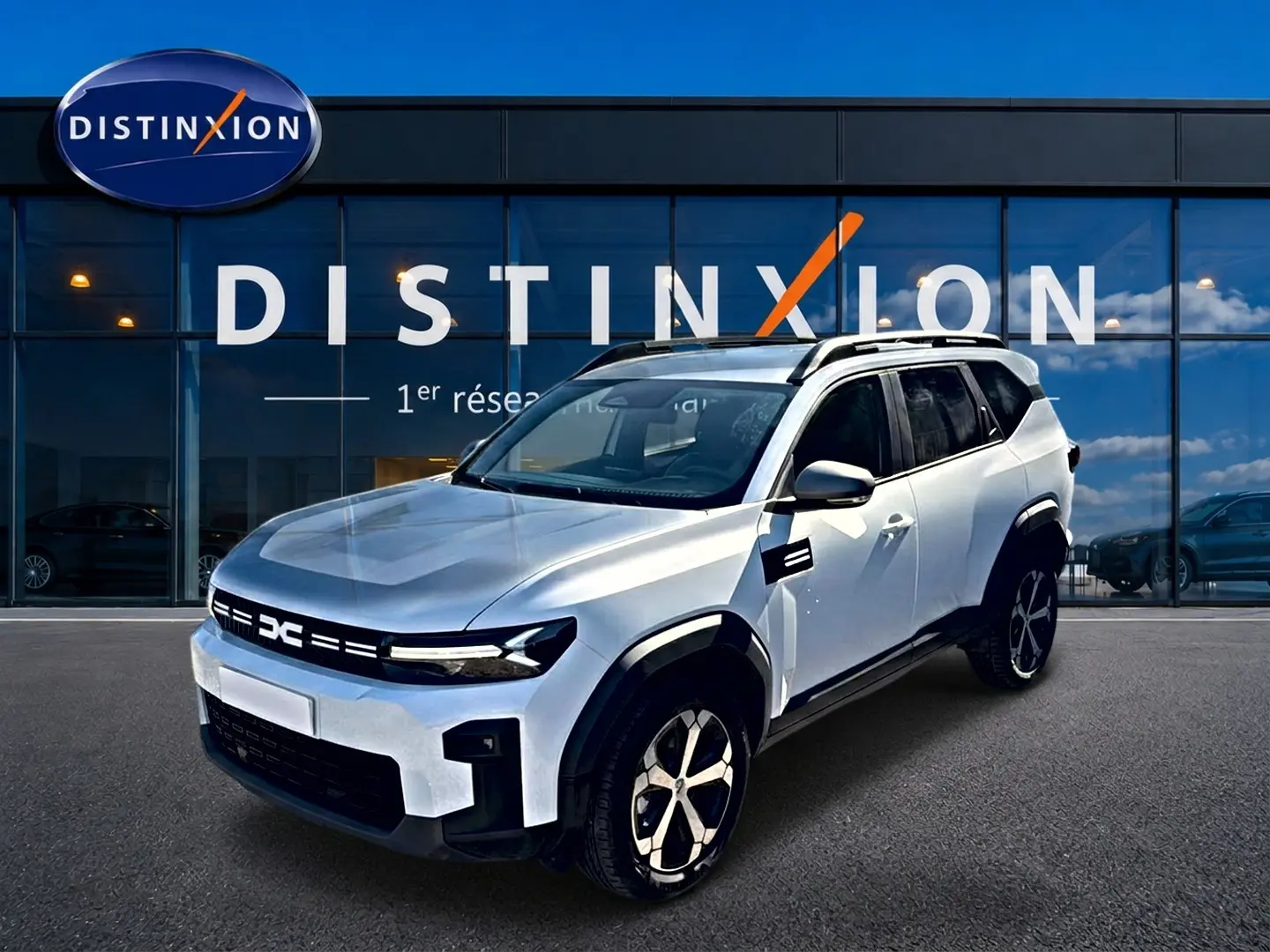 Dacia Bigster blanc en 3/4 avant droit, avec jantes alu et barres de toit visibles devant un showroom Distinxion.