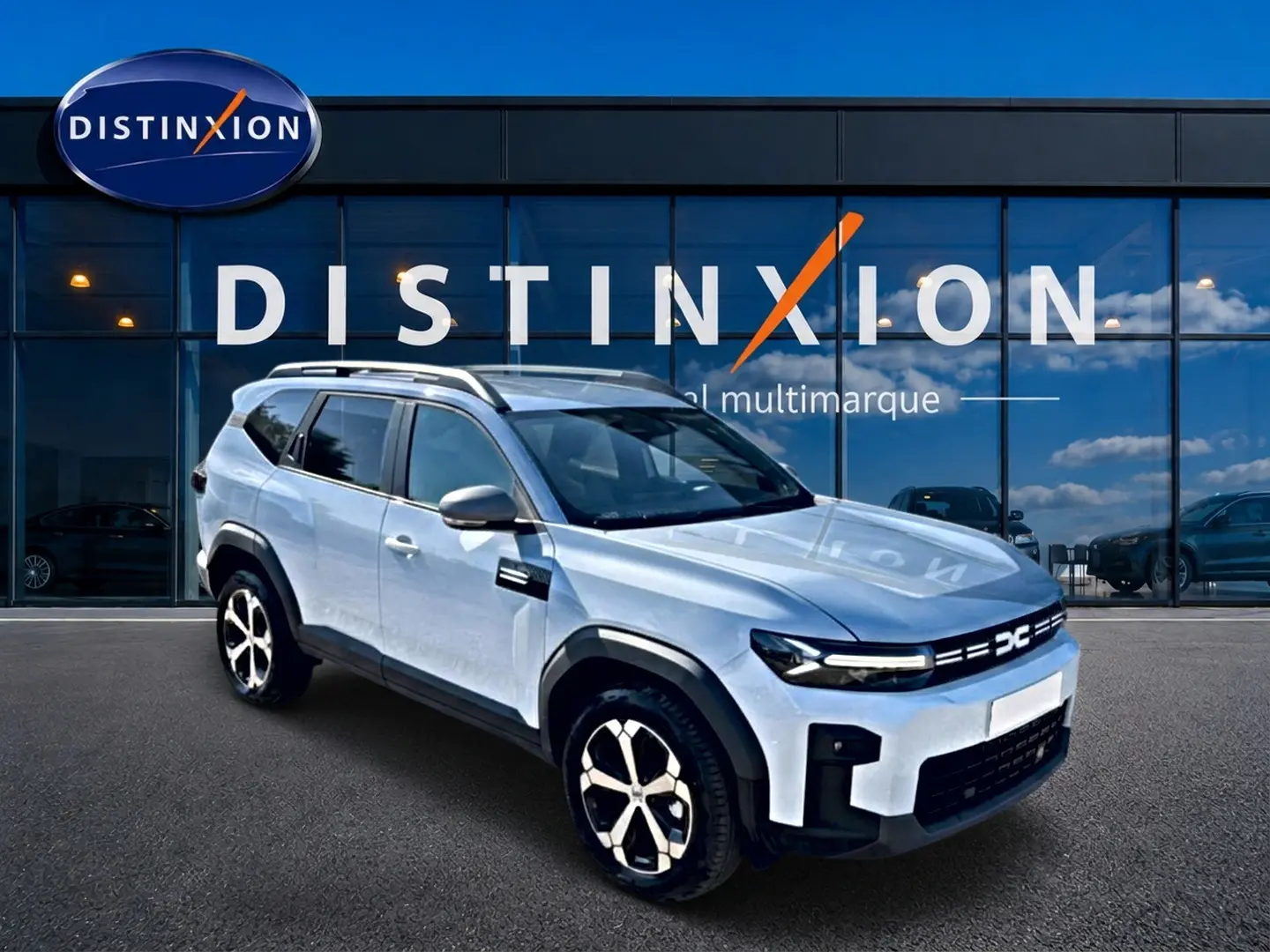 SUV Dacia Bigster blanc en 3/4 avant droit, avec barres de toit et jantes alu distinctives, devant une vitrine multimarque.