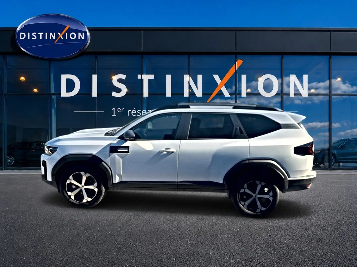 Profil droit du SUV blanc Dacia Bigster 2026 avec jantes alu et barres de toit devant une vitrine Distinxion.